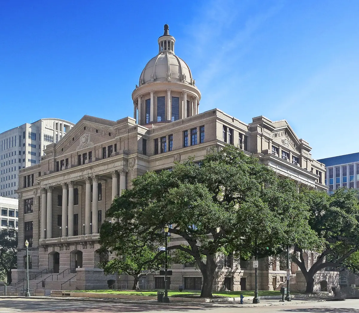 Restored beige stone courthouse with a copper dome and columns behind large oak trees in Houston